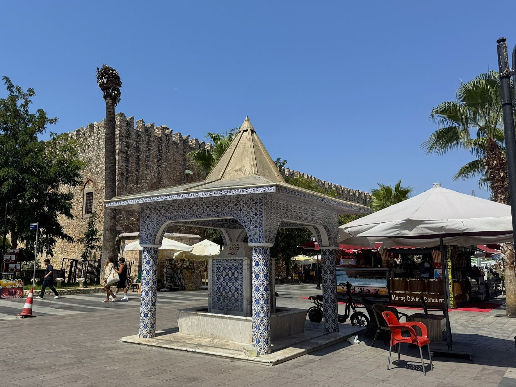 Ornate fountain in public square; stone building, blue and white tiles, food cart, blue sky.