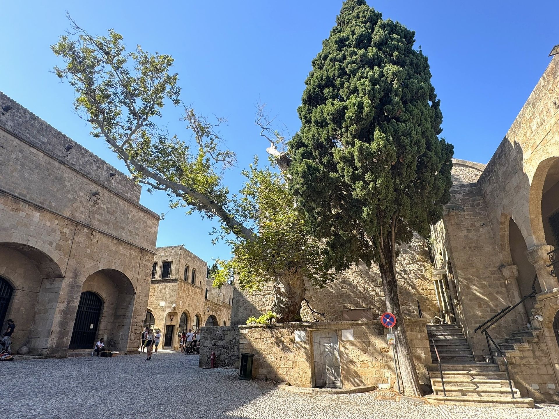 Courtyard in Rhodes, Greece, with stone buildings, trees, and people on a sunny day.