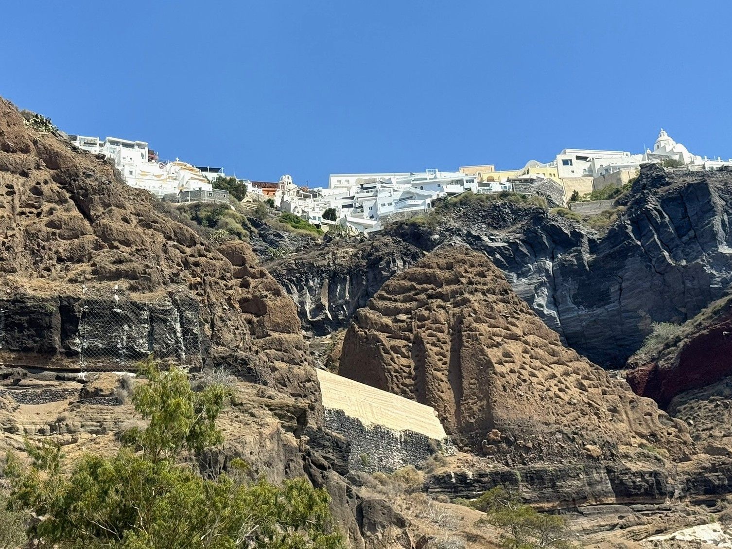 Cliffside town of white buildings overlooking textured volcanic rock and a clear blue sky in Santorini, Greece.
