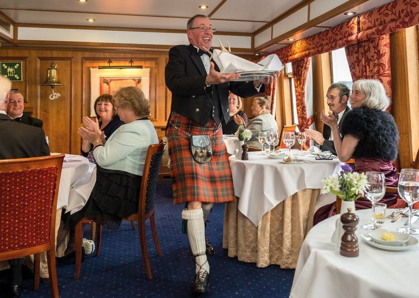 A man in a kilt is standing in front of a group of people sitting at tables addressing the haggis