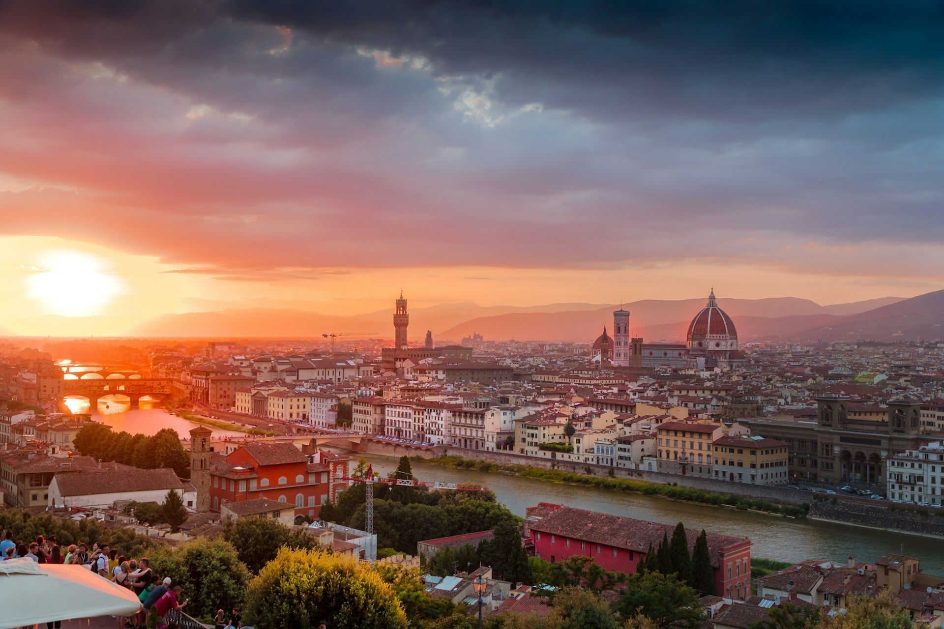 Experience the Festival dei Popoli, Florence on Azamara Sunset over Florence, Italy. Buildings, Arno River, Duomo, and Ponte Vecchio silhouetted against fiery sky.