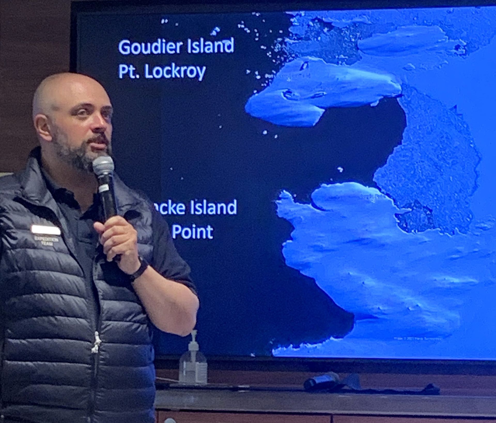 A man is holding a microphone in front of a screen that says goudier island pt lockroy