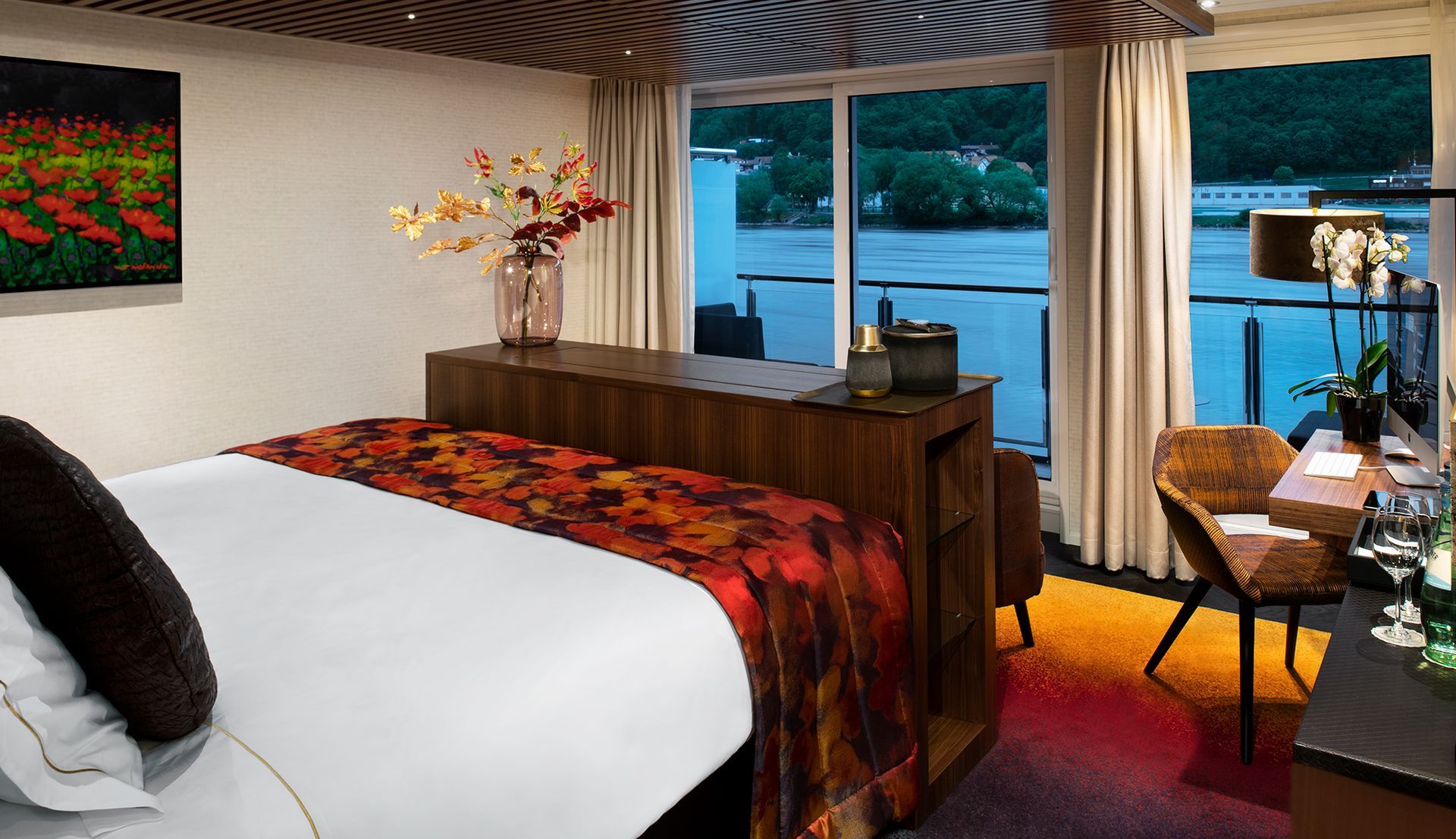 a river cruise ship cabin with a bed , desk , chair and a large window overlooking the water .