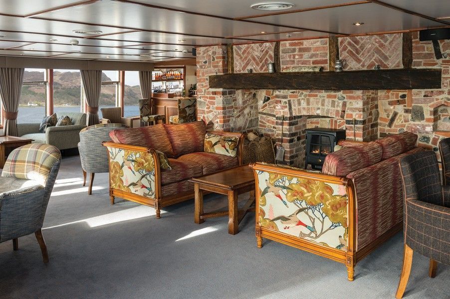 Tiree Lounge onboard Hebridean Princess Cozy lounge with floral sofas, brick fireplace, and a scenic view of the water and mountains.