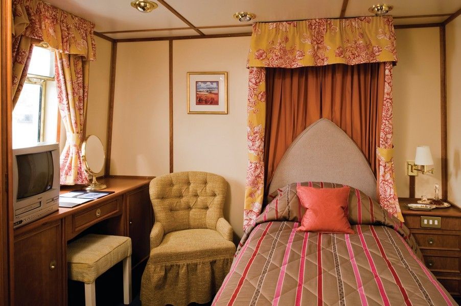 Delightful accommodation on board Hebridean Princess Cozy bedroom with a small bed, desk, armchair, and window with curtains. Earth-tone color scheme.