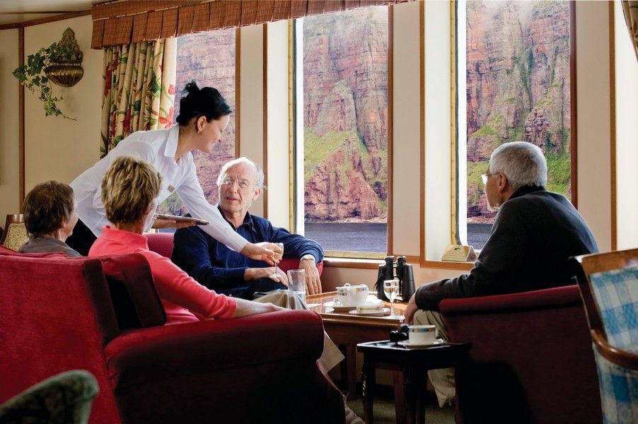Exceptional service onboard A waitress serves a drink to an elderly man, seated with others in a cabin overlooking a scenic landscape.