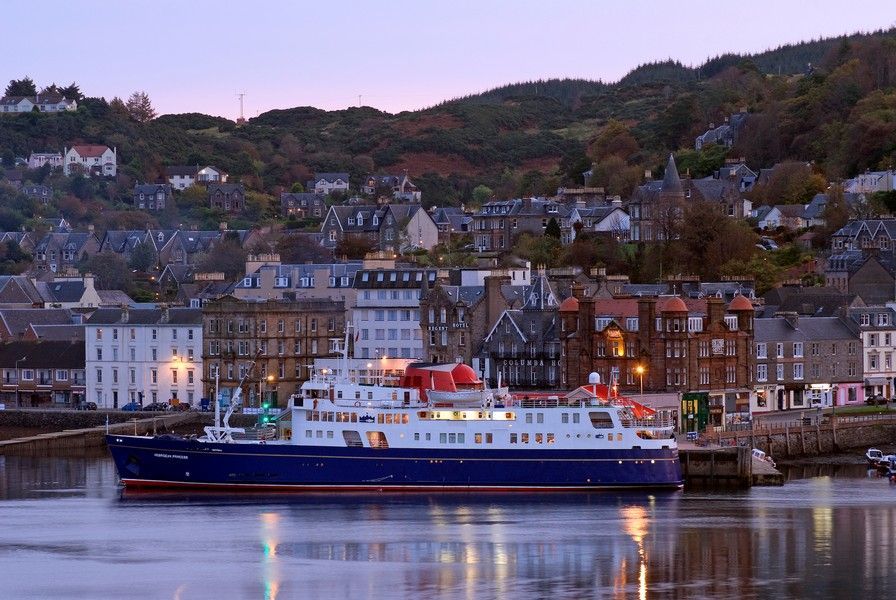 Hebridean Princess: A truly Unique Ship Large blue and white ship docked in a harbor town at dusk, buildings with lit windows in the background.