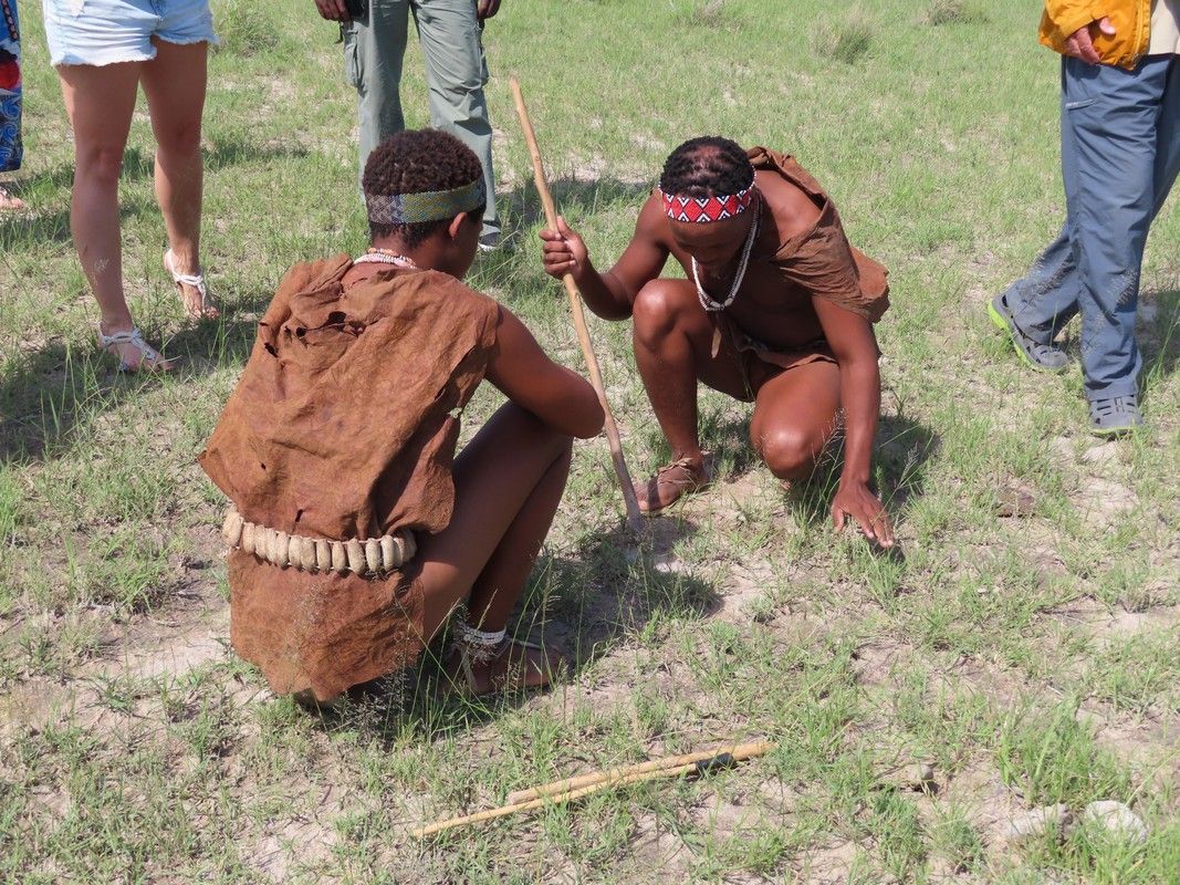 Two men are squatting down in the grass holding sticks.