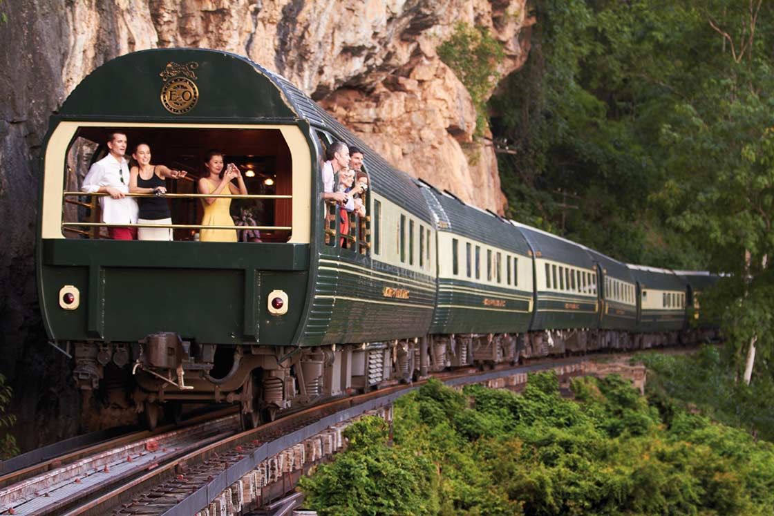 Green luxury train on tracks, passing by a rocky cliffside and lush greenery. Passengers wave from open windows.