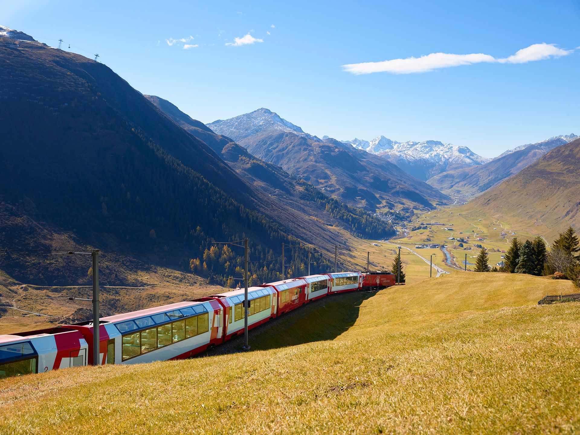 Train traveling through a mountain valley with red and white cars, golden grass, and blue sky.