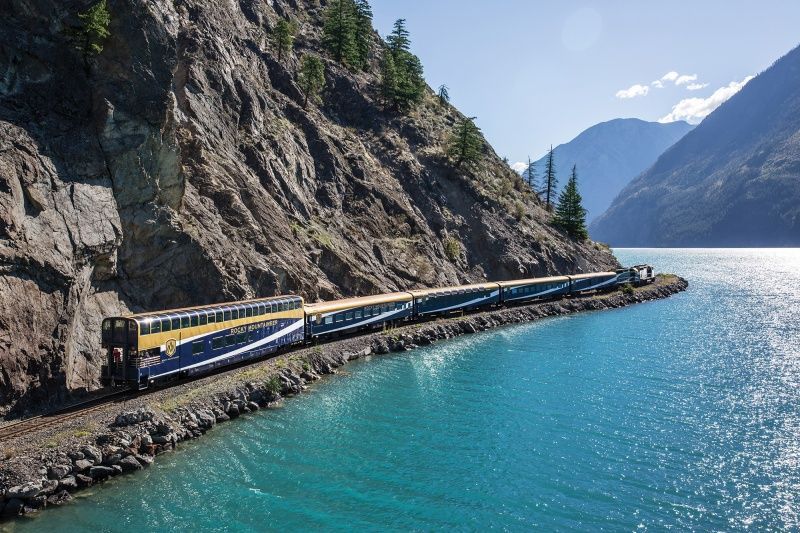 Train travels along a rocky cliffside next to a turquoise lake, mountains in the background.