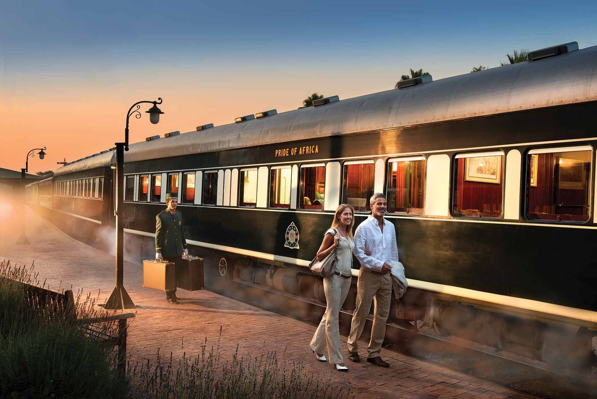 Couple walking towards a luxury train at sunset, with attendant nearby.
