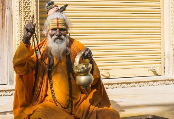 A man with a beard and a yellow robe is sitting on the ground holding a bell in India