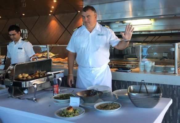 a cruise ship officer is standing in front of a buffet table with plates of food on it  on Azamara Quest