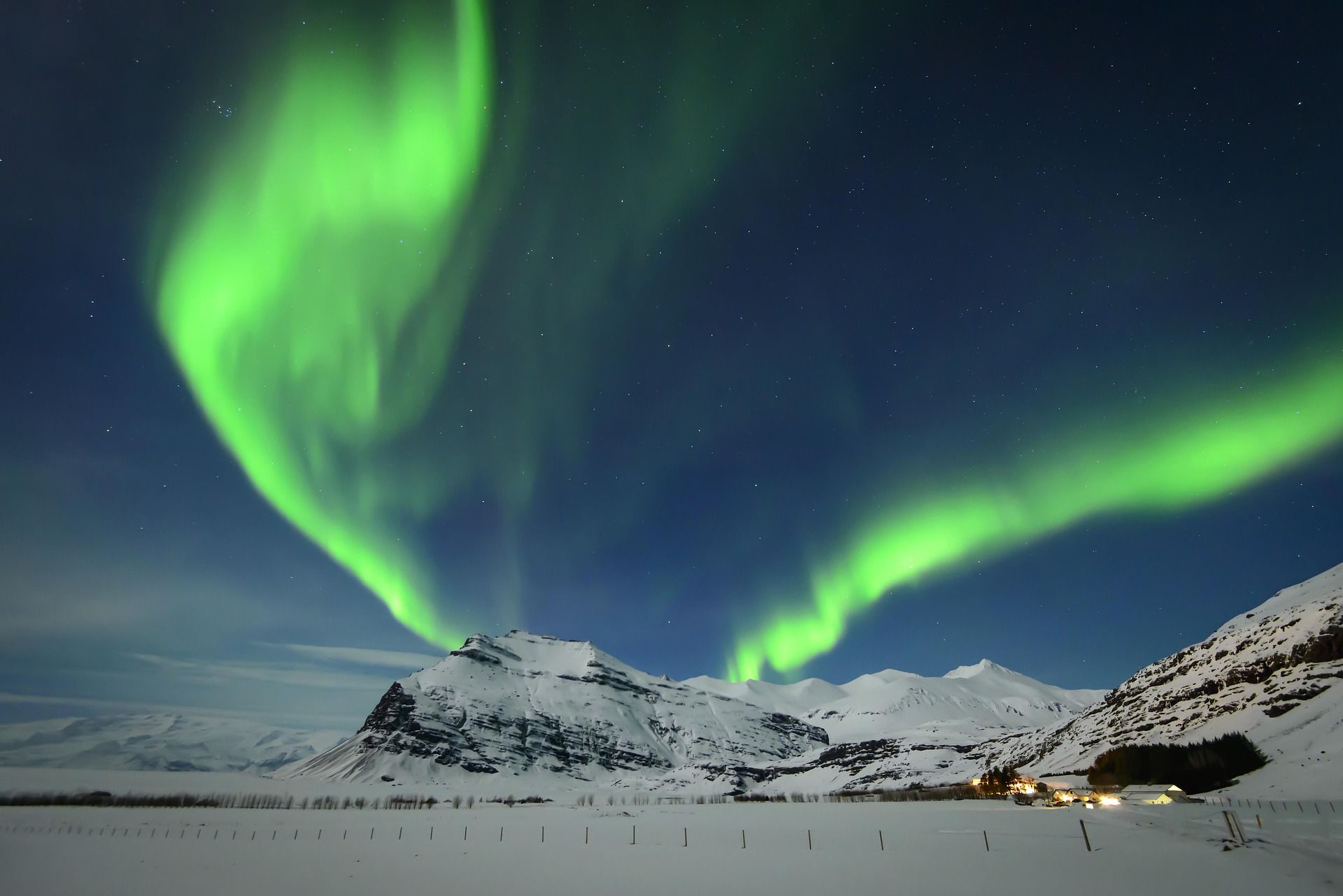 Green aurora borealis dances above snow-covered mountains and a small village under a dark blue sky 