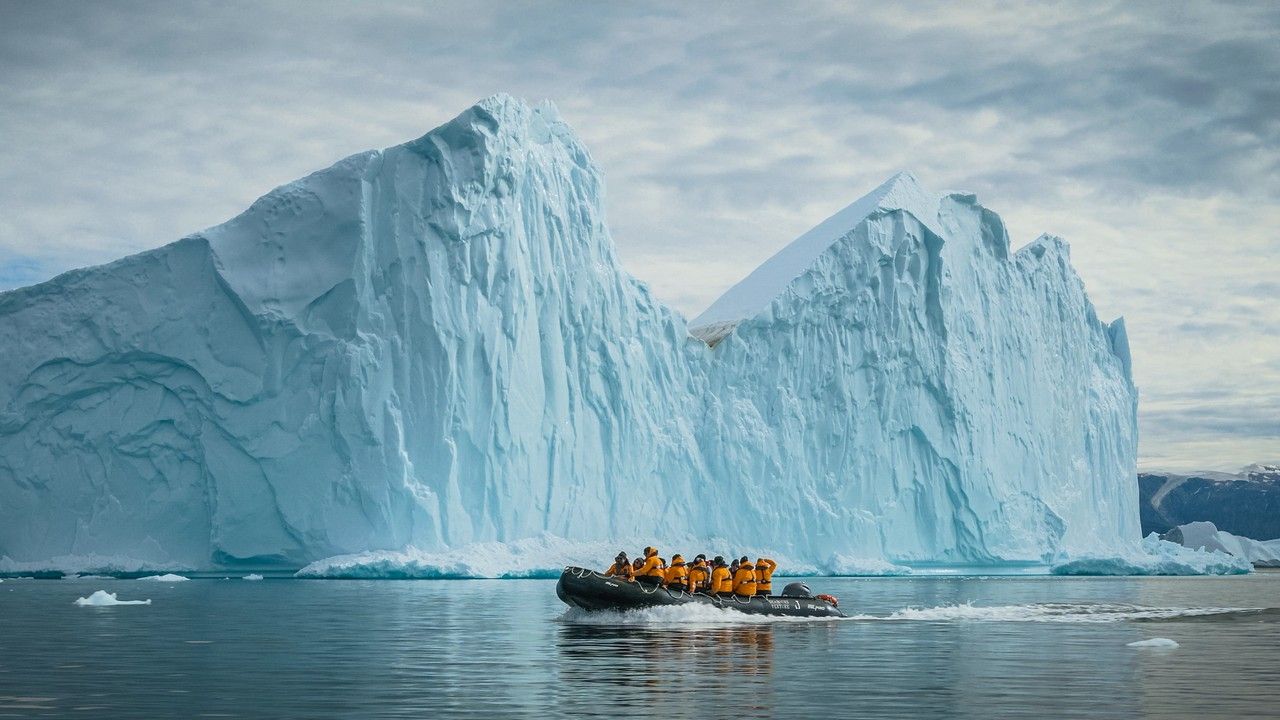 Seabourn Expedition guests on a zodiac tour A small boat with people in orange jackets sails near a massive iceberg in an arctic setting.