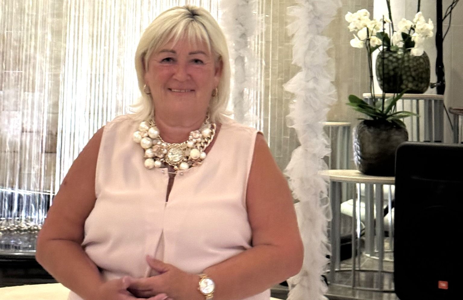 Woman with blonde hair in a pink top, wearing a statement necklace, smiling. Indoors.
