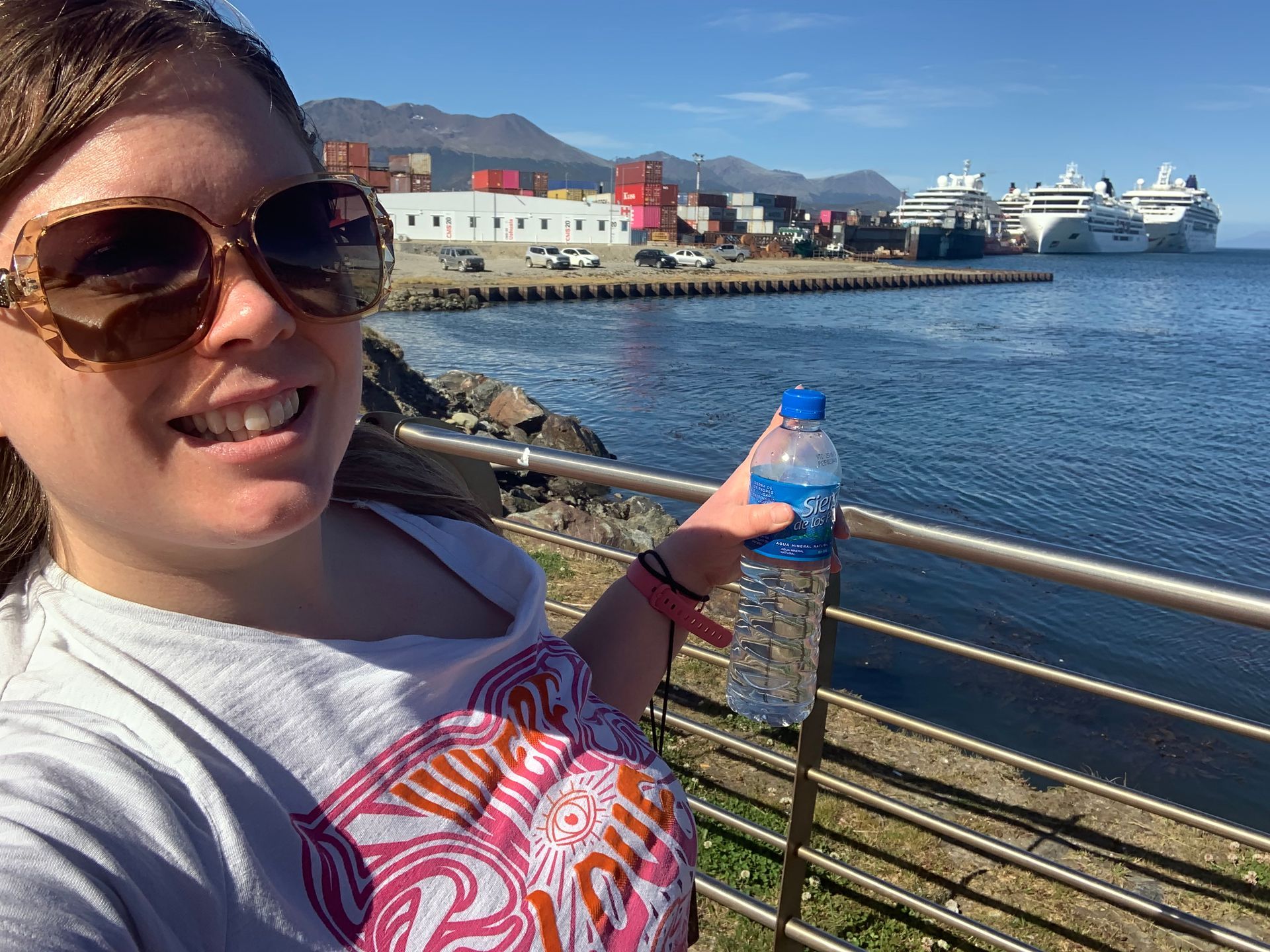 A woman wearing sunglasses is holding a bottle of water in front of a body of water pointing at ships in Ushuaia