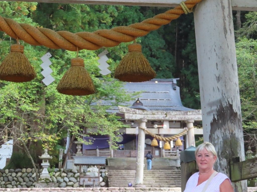 Woman poses in front of a Japanese shrine with a rope decoration. Trees surround the temple.