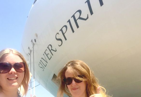 Two women wearing sunglasses are standing in front of a silver spirit airplane.