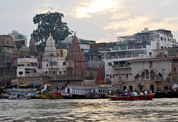 A group of people are riding boats down a river in front of a city in India