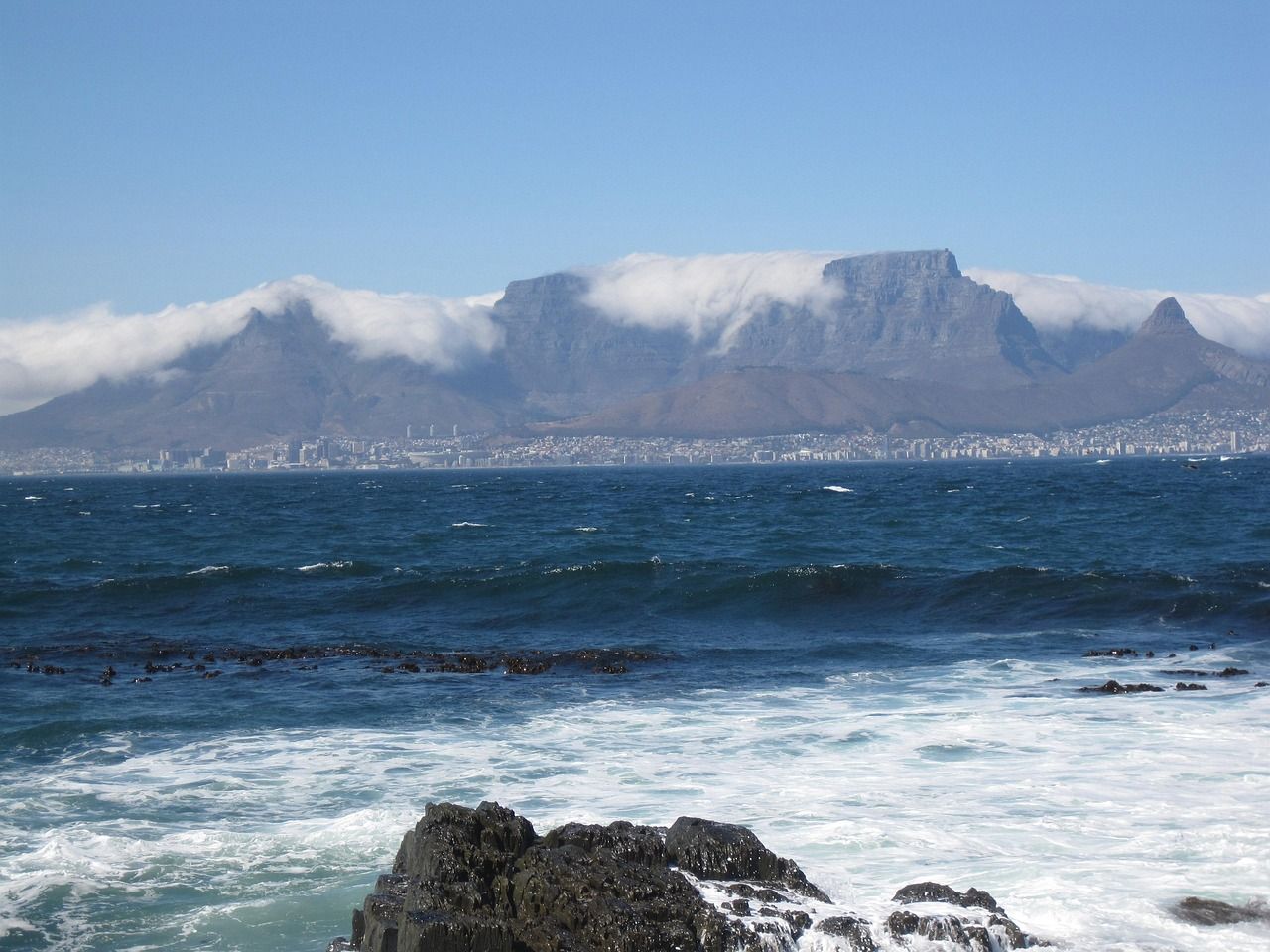 Table Mountain covered in clouds, seen from the ocean with waves crashing on foreground rocks; clear blue sky.