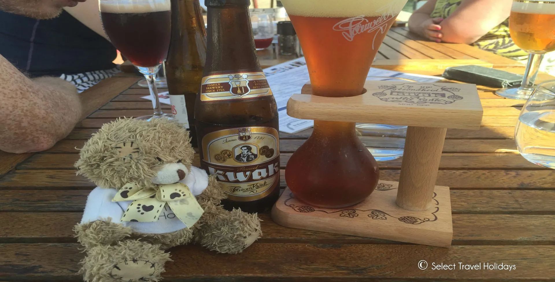 Teddy bear next to a bottle and glass of kwak beer on a wooden table, outdoors.