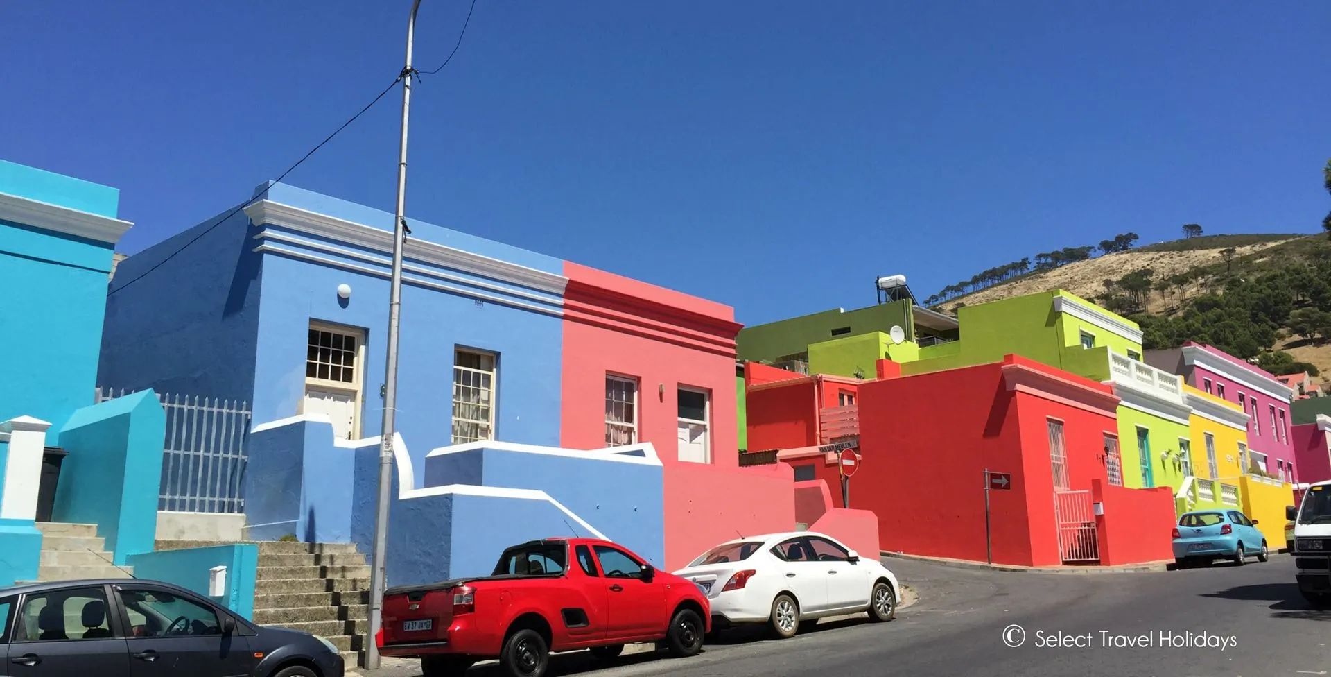 Colorful houses on a street in Bo-Kaap, Cape Town, South Africa. Blue sky, cars parked on the road, and a hill in the background.