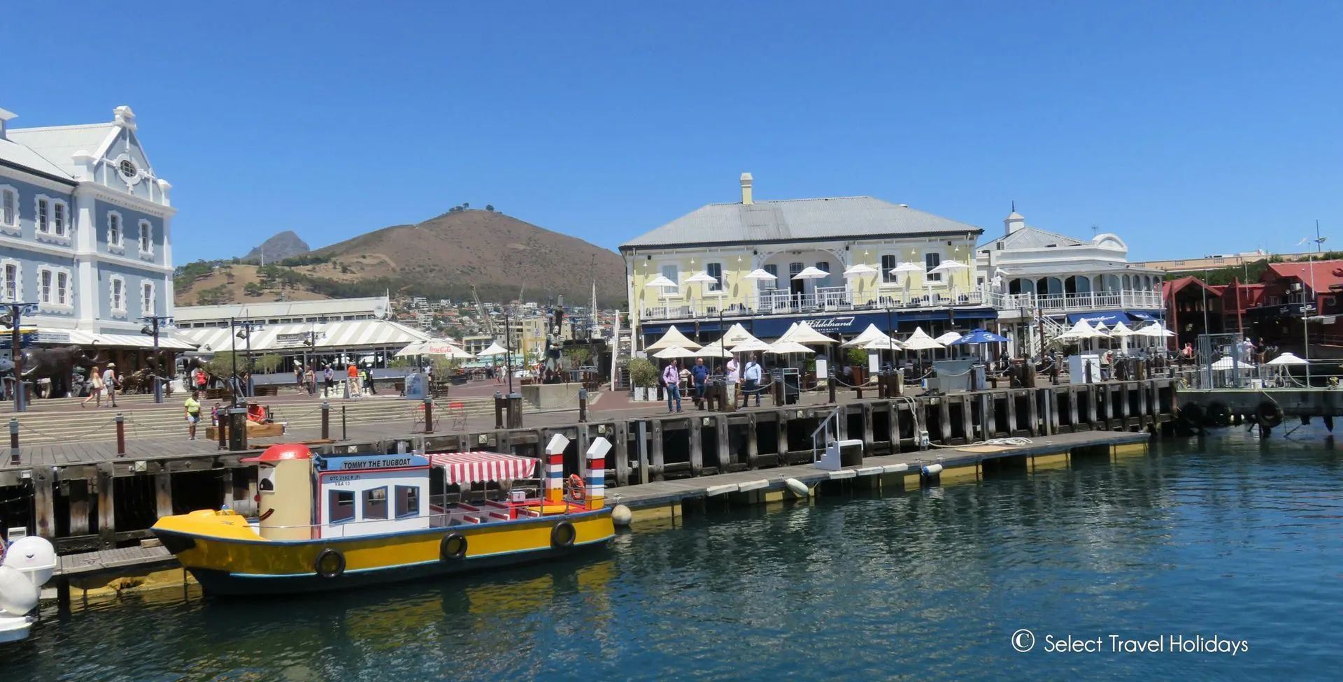 Waterfront scene in Cape Town, South Africa, with colorful buildings, a boat on the water, and people walking on the pier on a sunny day.