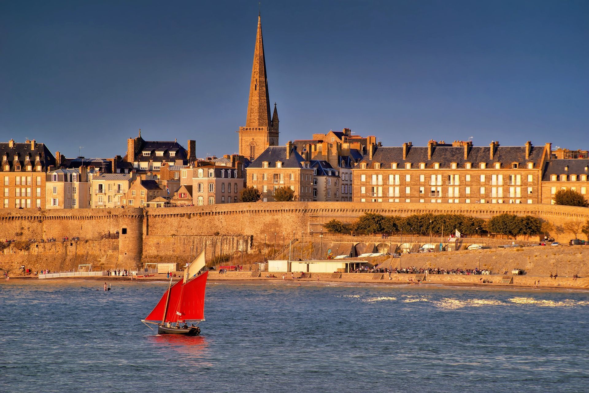 A red sailboat is floating in the ocean in front of a city.