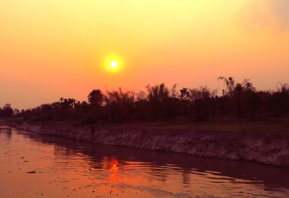 The sun is setting over the Brahmaputra river with trees in the background.