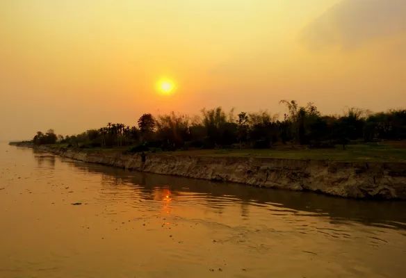 The sun is setting over the Brahmaputra river with trees on the shore.