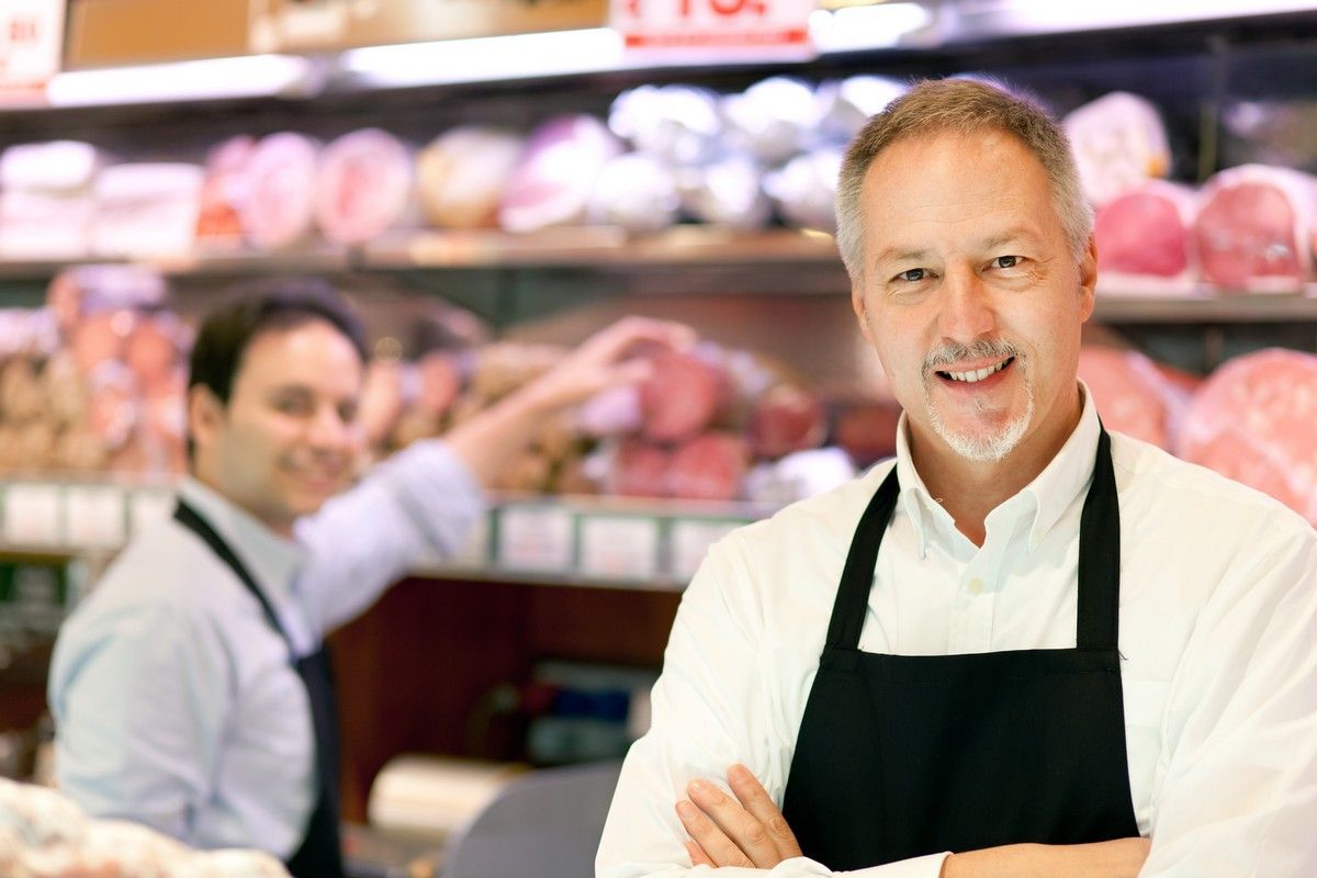 A man in an apron is standing in a grocery store with his arms crossed