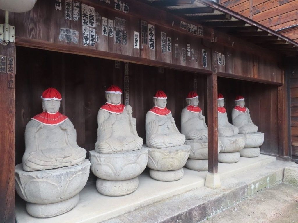 Six seated stone statues with red hats and collars, in a wooden structure.