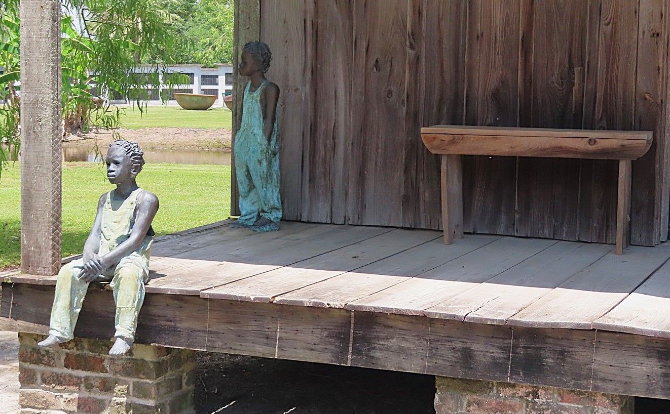 A statue of a boy sitting on a porch next to a bench.