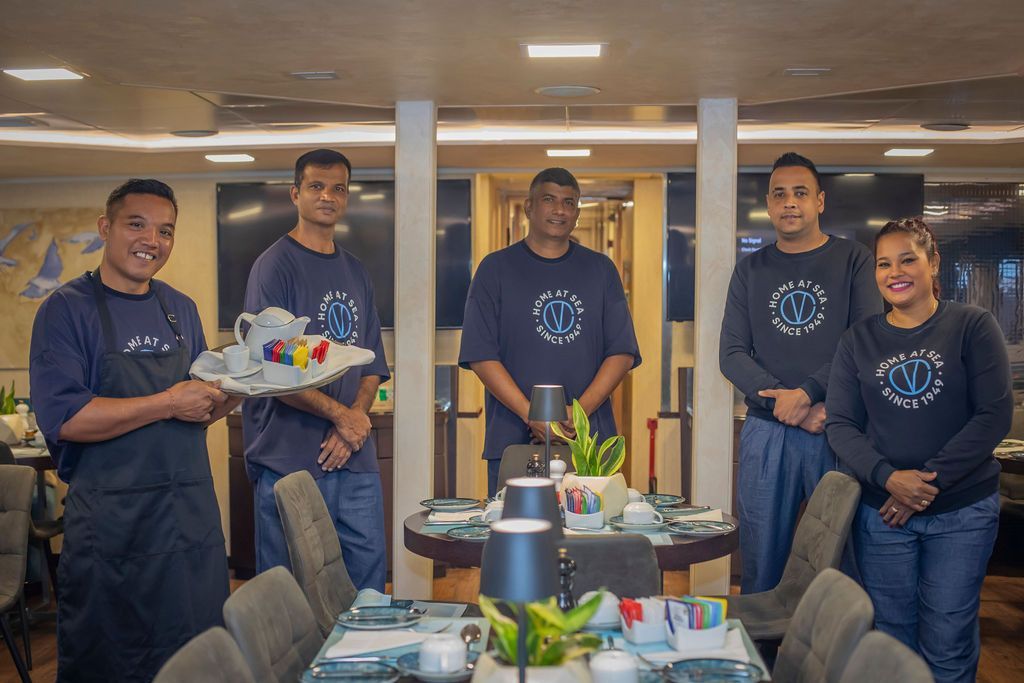 A team of five people poses in a dining room; one holds a tray with tea set, table set for dining.