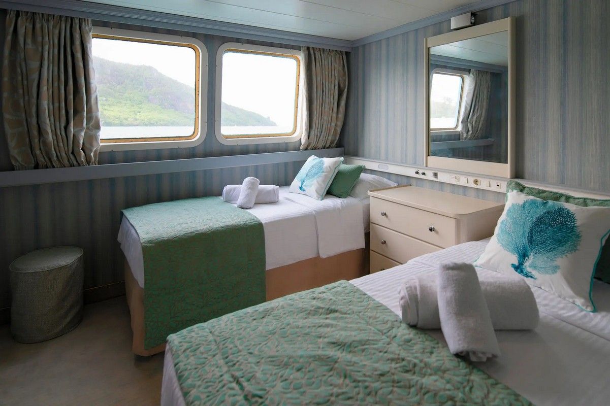 Twin beds in a stateroom with a window view, white and green linens, mirror, and striped walls.