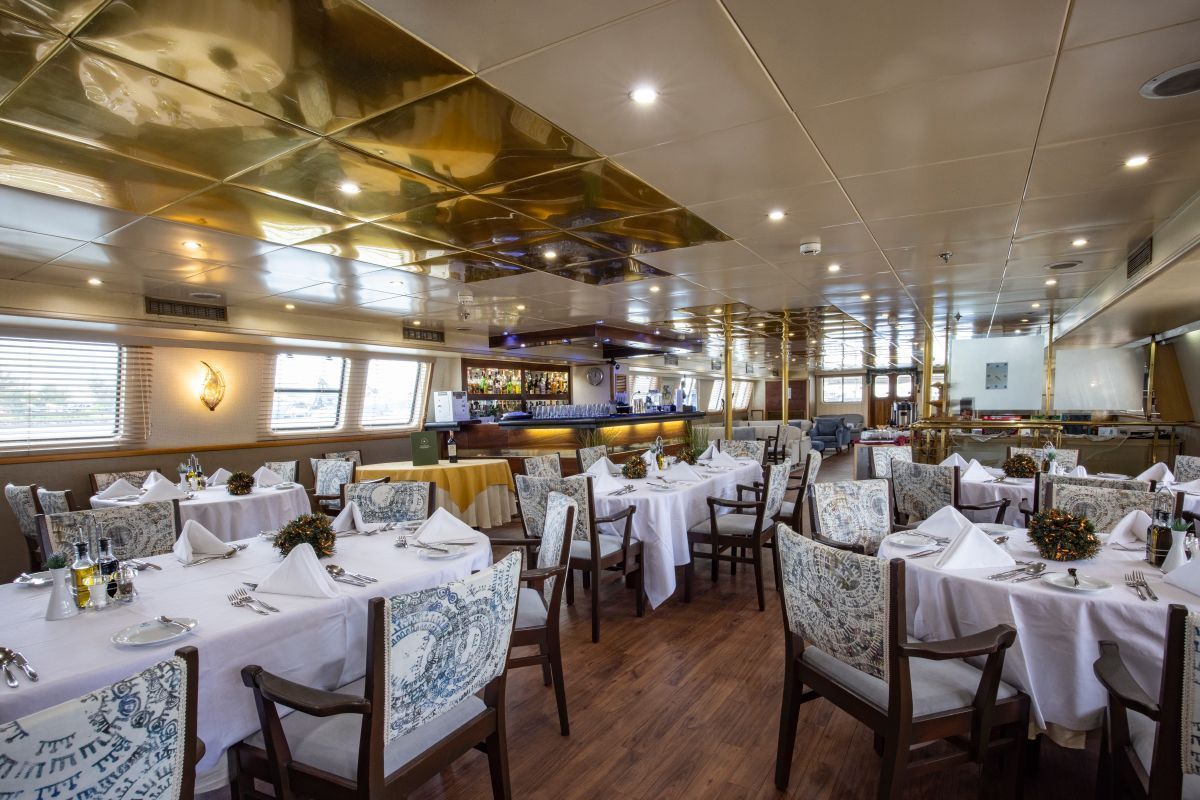 Restaurant onboard the ship Formal dining room on a boat with round tables, white tablecloths, and ornate chairs; gold ceiling.