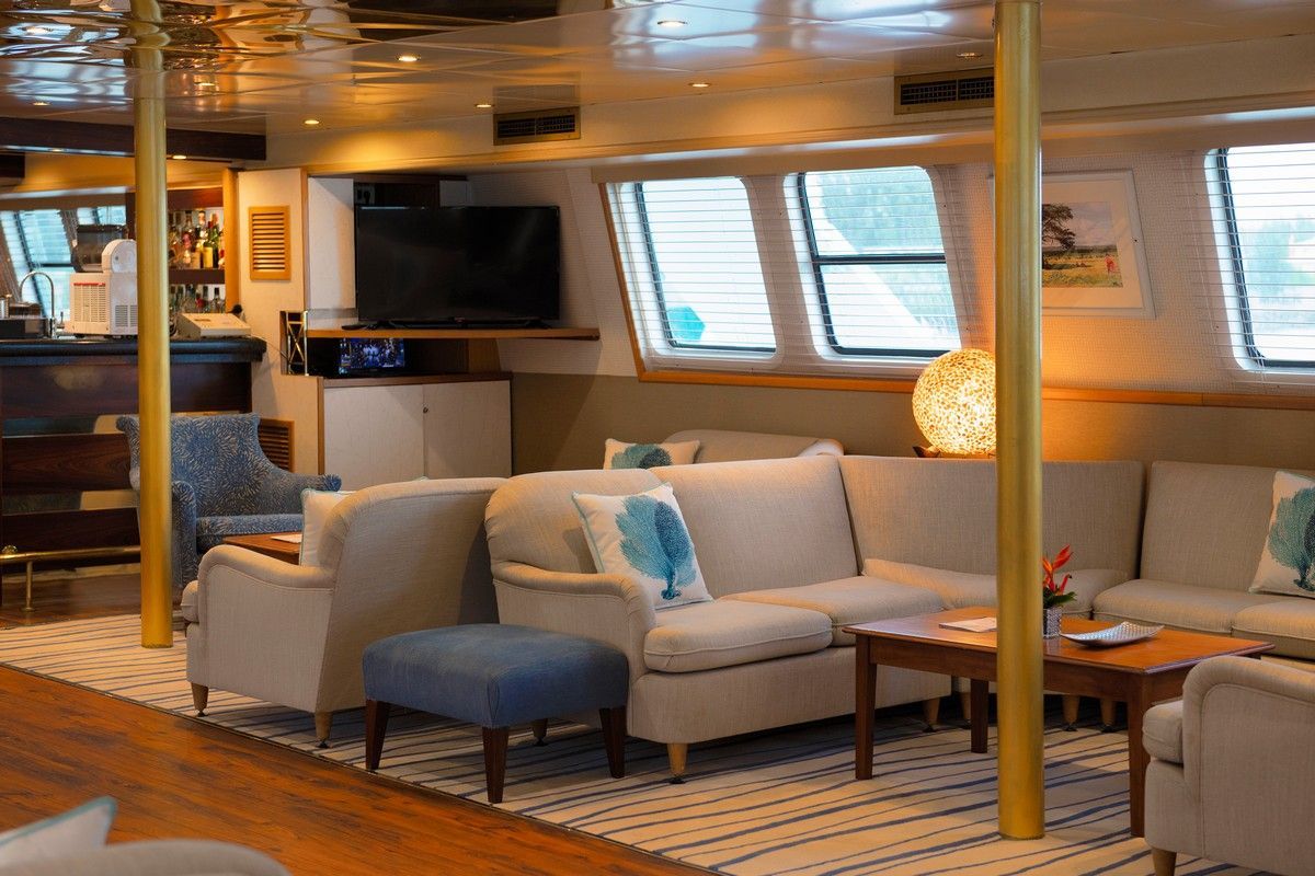 Lounge area on a ship with beige sofas, blue accents, a TV, and a bar.