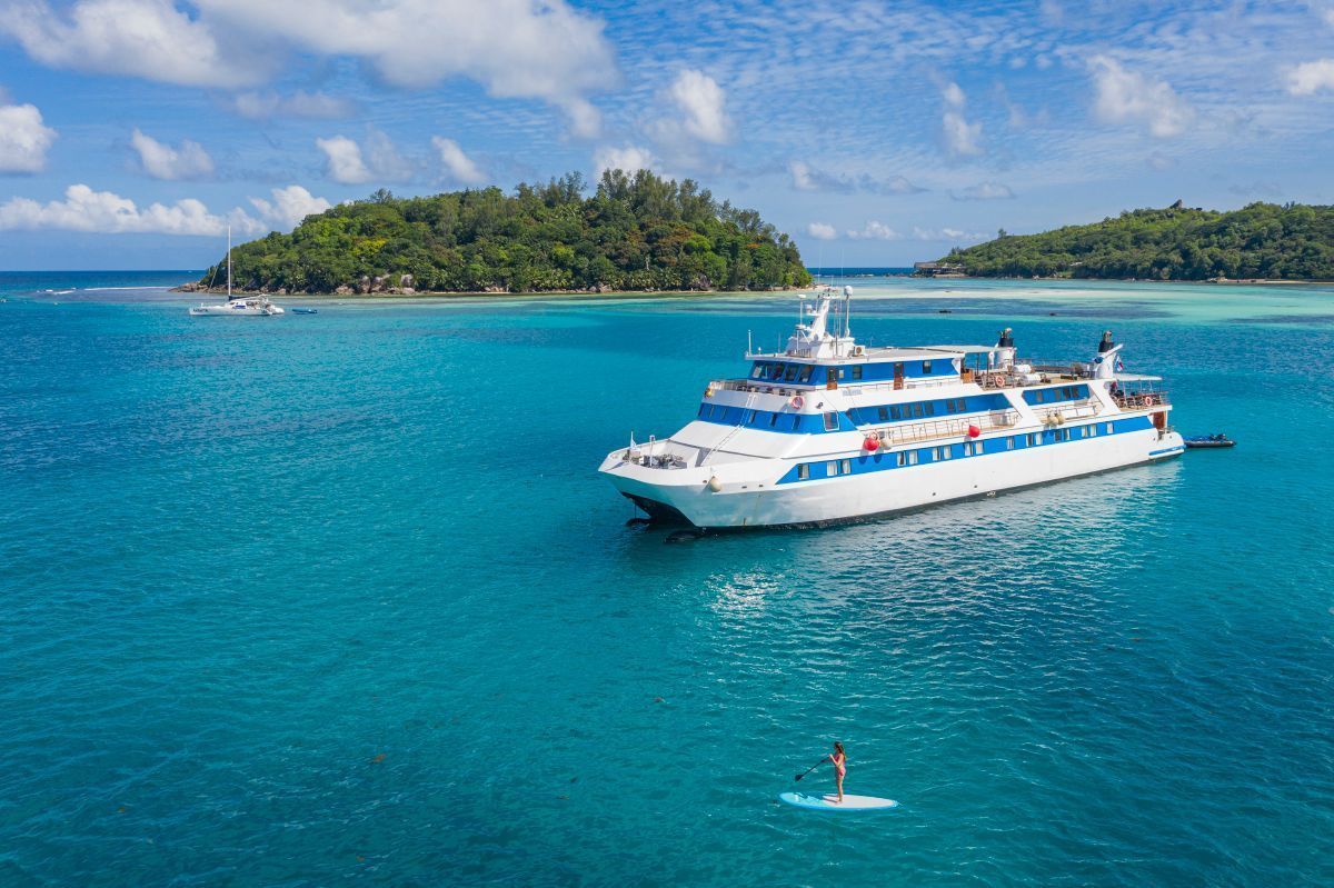 Large white and blue boat on turquoise water, a person paddleboards. Islands in background.