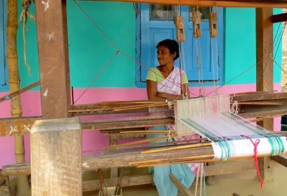 A woman is sitting at a loom in front of a colorful building in Assam, India