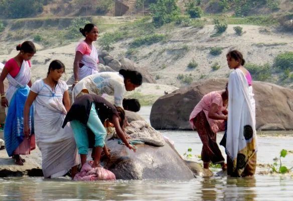 A group of people are standing in the water near a large rock washing clothes on the Brahmaputra river