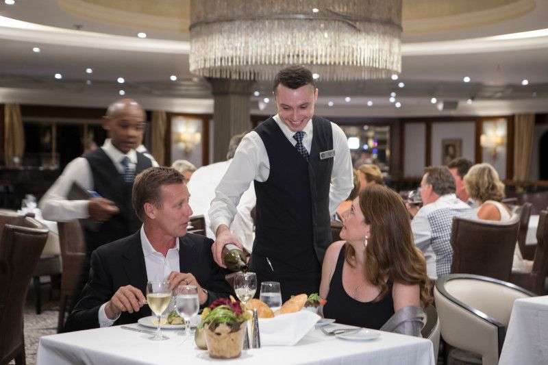 A man is pouring wine into a glass for a woman in a restaurant whilst dining on Azamara