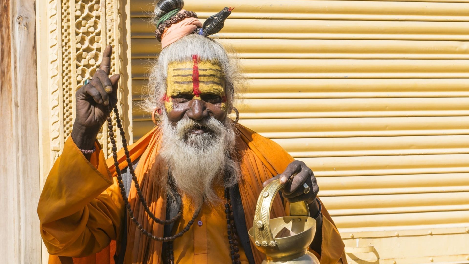 a man with long hair and a beard is standing in front of a building holding a bell at the Kumbh Mela, India