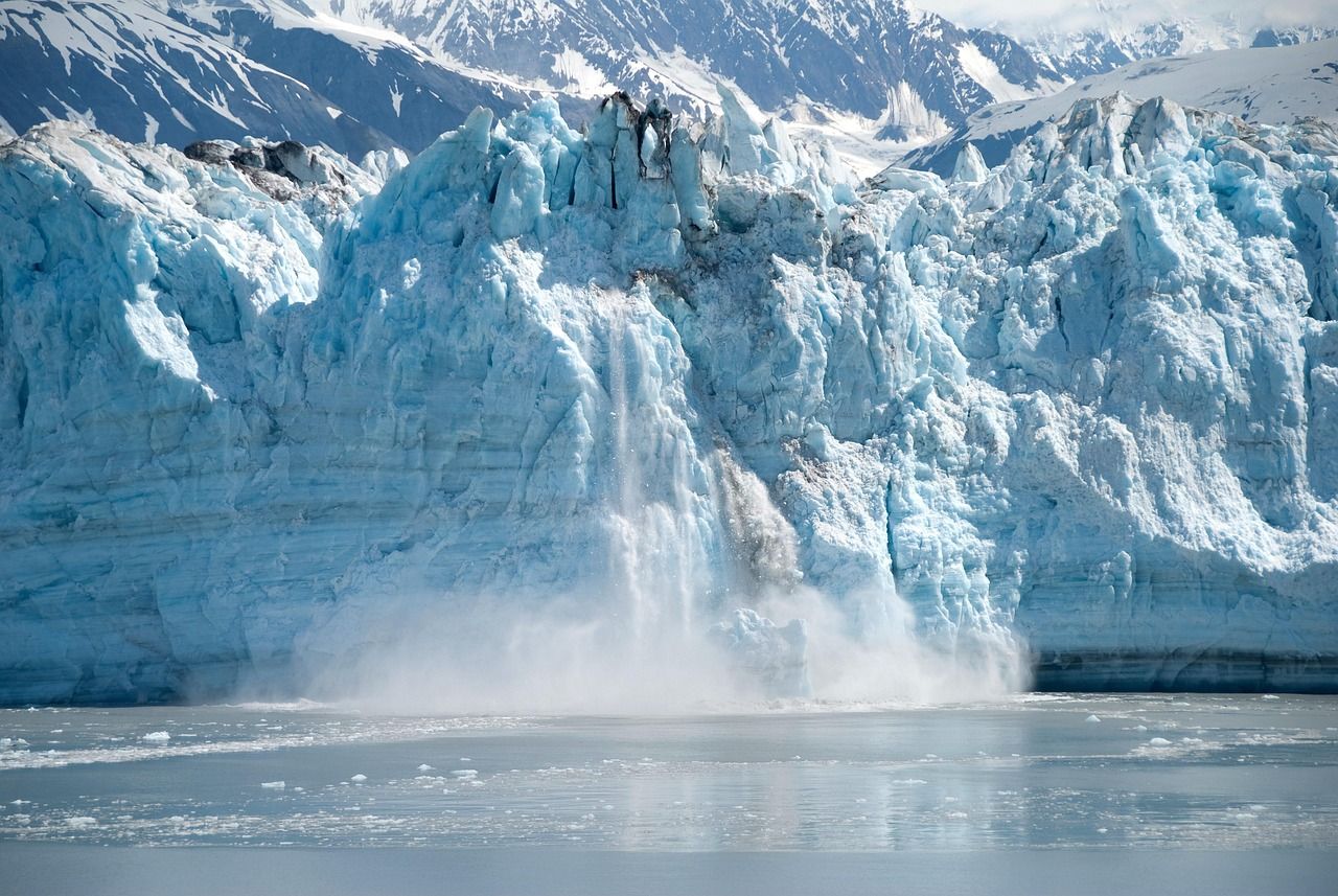 A large iceberg is breaking into a body of water with mountains in the background.