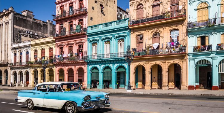 A blue and white car is driving down a street in front of a row of colorful buildings in Cuba