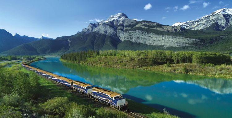 A train is going down the tracks next to a river with mountains in the background.