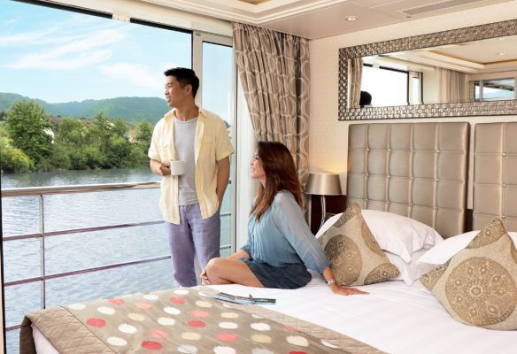 a man is standing next to a woman sitting on a bed in a  balcony cabin on AmaKristina river cruise ship