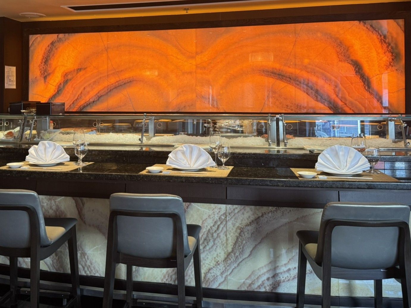 Sushi bar with three seats, folded napkins, and an orange, abstract mural.