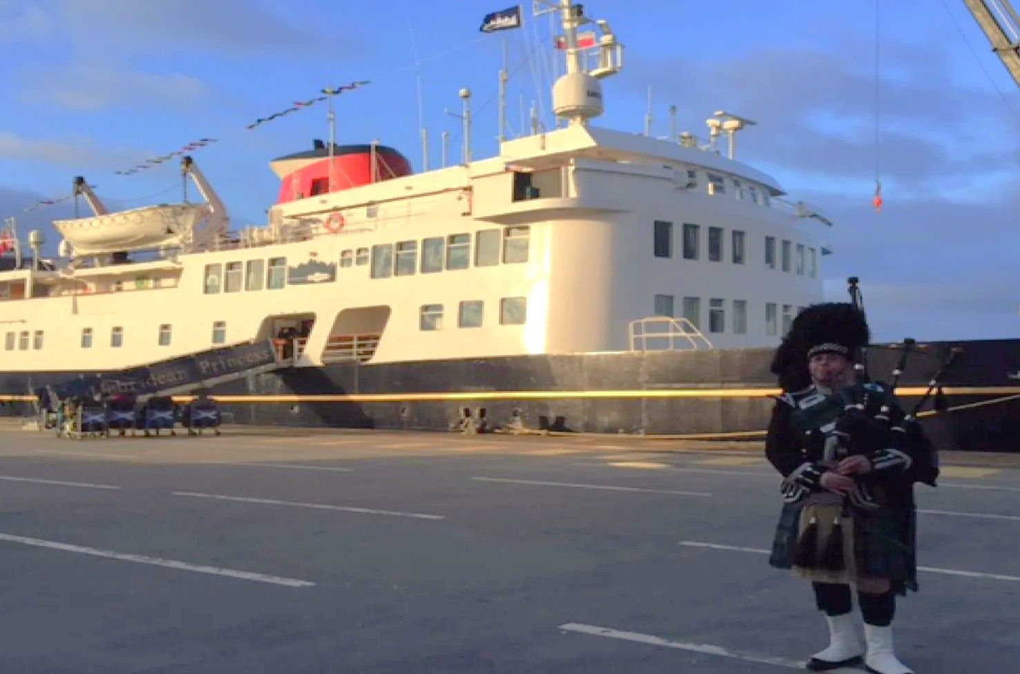 A warm welcome in Greenock A bagpiper in kilt stands on dock near a cruise ship, likely welcoming passengers.